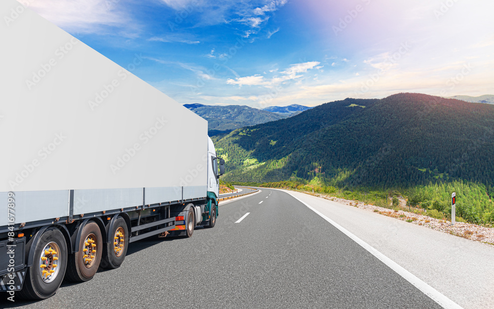 A white truck transports cargo along the highway against the backdrop of a mountain landscape.
