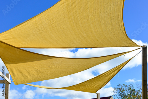 Multiple large summer nylon patio sun shade umbrellas, sunny yellow color with wooden supports. The sun is shining through the umbrellas. The background is a bright blue sky, trees, and a building.