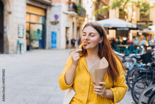 Carefree woman eating traditional Churros, a fried pastry with chocolate on a city street. Positive cheerful female student posing outdoors. Food concept
