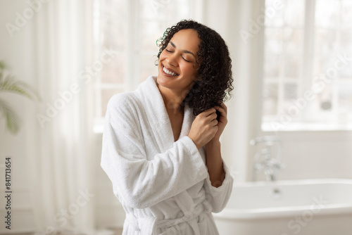 Haircare concept. Pretty black lady in bathrobe touching her hair after morning bath, standing in bathroom, enjoying beauty routine caring for herself