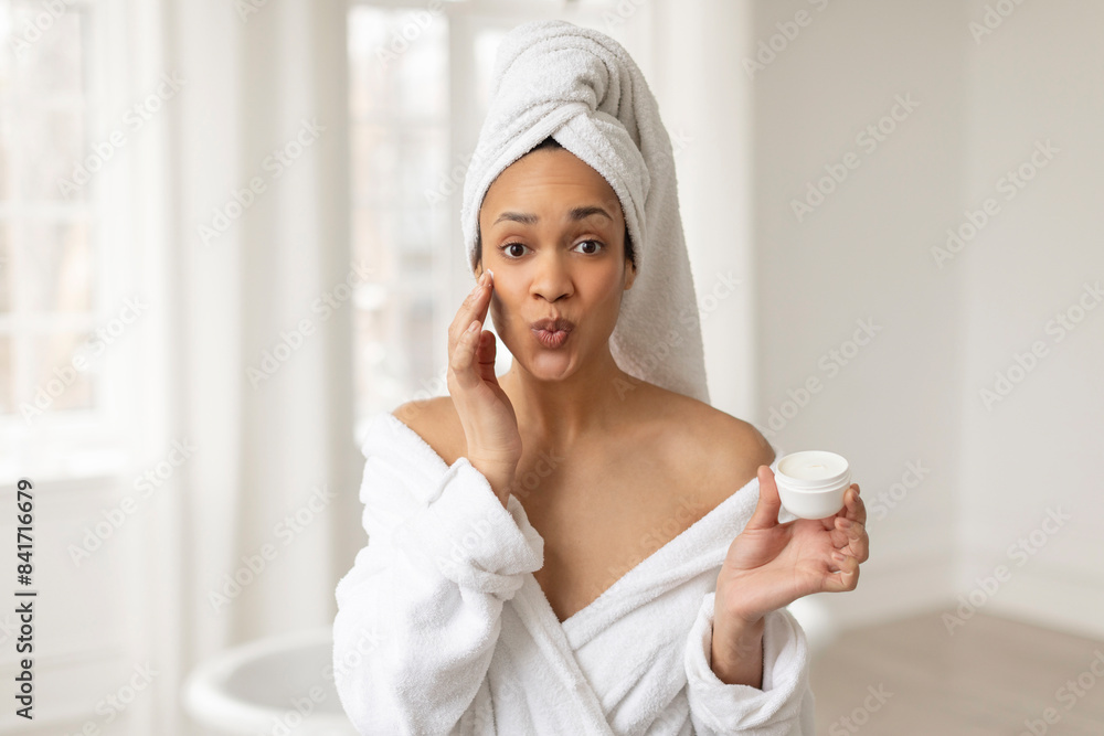 Facial skincare concept. Joyful black woman applying moisturizer cream on face and pouting lips. Skin care beauty product Facial skincare concept. Joyful black woman applying moisturizer cream on face and pouting lips. Skin care beauty product