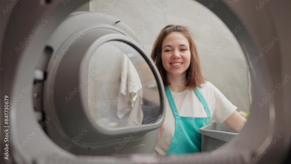 A happy brunette girl in a white T-shirt and a blue apron took out clean things from the washing machine and closes its door while cleaning the apartment on call A happy brunette girl in a white T-shirt and a blue apron took out clean things from the washing machine and closes its door while cleaning the apartment on call