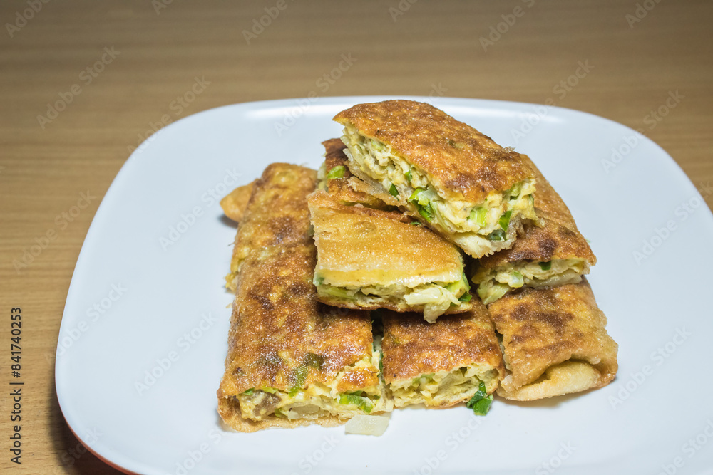 Indonesian food: Martabak Telur (omelette murtabak/mutabbak/matabbak/mutabbaq), which is a folded omelette pancake with bits of vegetables, on top of white rectangular plate, on top of wooden table Indonesian food: Martabak Telur (omelette murtabak/mutabbak/matabbak/mutabbaq), which is a folded omelette pancake with bits of vegetables, on top of white rectangular plate, on top of wooden table