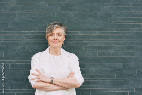 Portrait of 55 - 60 year old woman with grey hair, wearing white jacket, posing on grey background