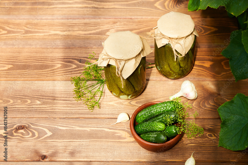 Pickled cucumbers in jars, fresh cucumbers on bowl, dill, garlic on a wooden background. Home canning