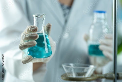 A scientist wearing a lab coat and gloves holds a conical flask containing a blue liquid photo