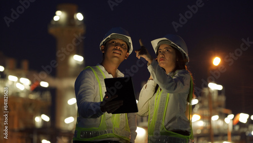 Two engineers wearing helmets and reflective vests use a tablet to discuss operations at a brightly lit industrial site during nighttime.