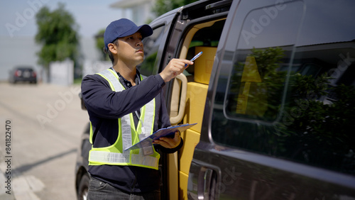 A delivery worker in a cap and reflective vest uses a clipboard to check packages stacked inside a van parked outdoors.