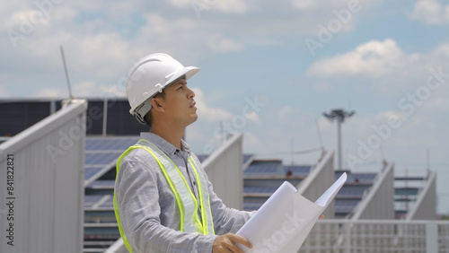 An engineer in a hard hat and safety vest reviews solar project plans on a tablet, standing at a site with solar panels.