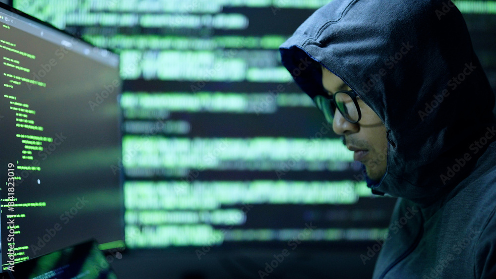 A person wearing a hoodie and glasses closely examines code on multiple screens, suggesting hacking or programming activity in a dark room. A person wearing a hoodie and glasses closely examines code on multiple screens, suggesting hacking or programming activity in a dark room.