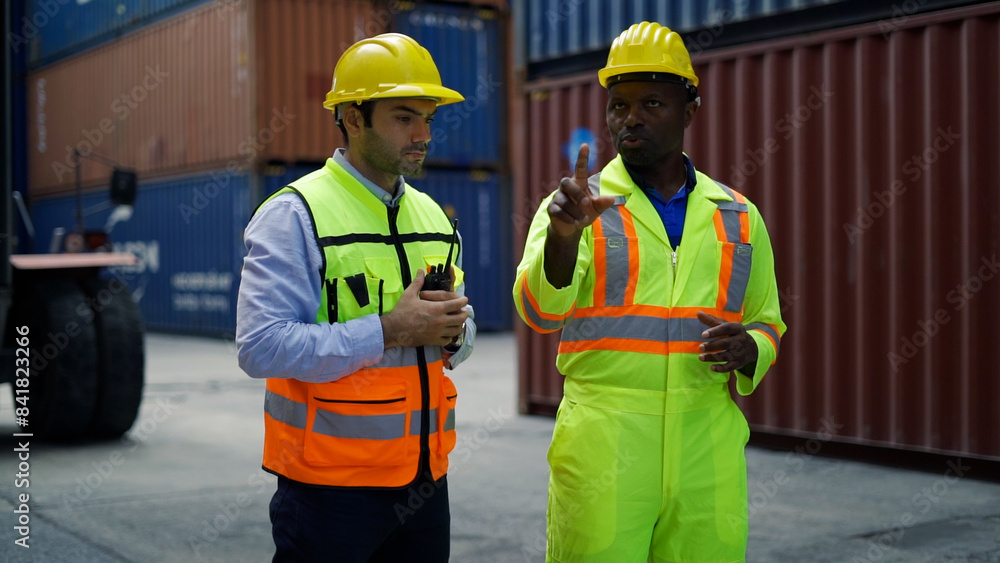 Two workers wearing hard hats and safety vests discuss logistics and operations at a shipping container yard with stacked containers in the background. Two workers wearing hard hats and safety vests discuss logistics and operations at a shipping container yard with stacked containers in the background.
