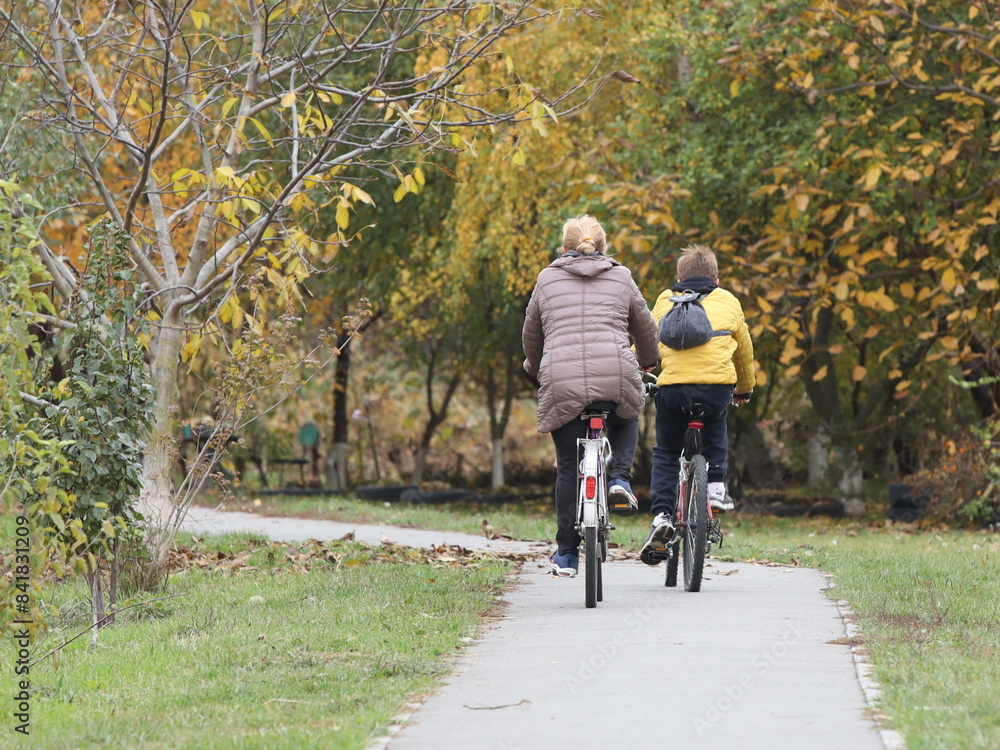 Fototapeta premium Mom and son ride bicycles along the alley of an autumn park. Healthy and active lifestyle in raising the younger generation. Active recreation in nature. Traveling on two wheels