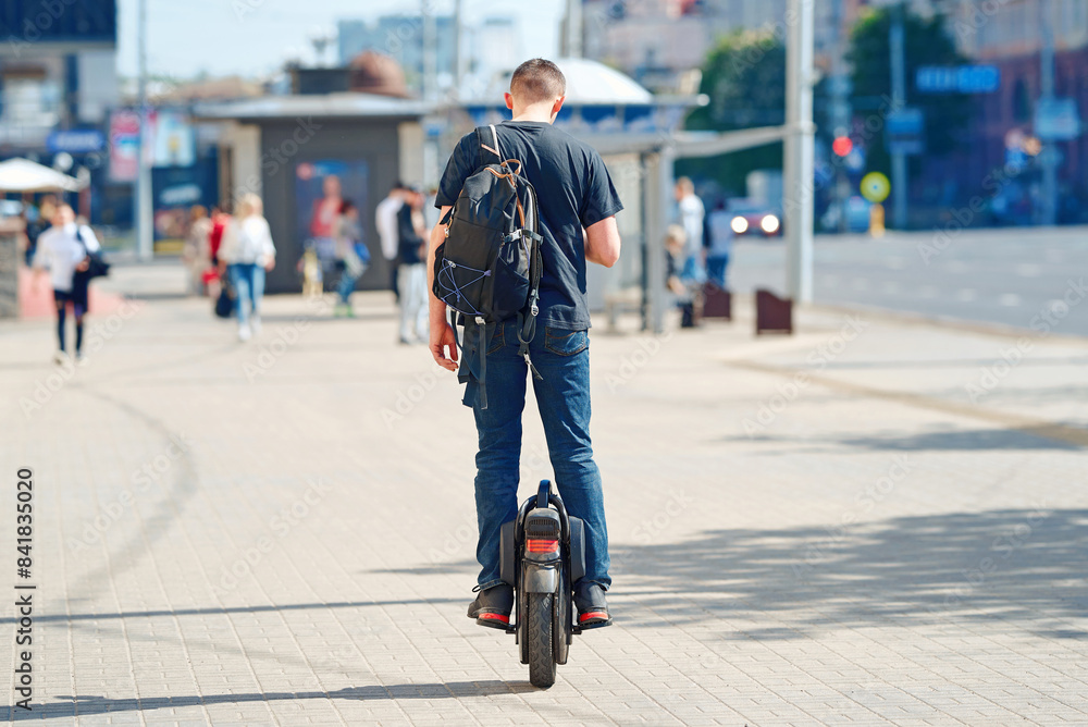 Obraz premium Man with backpack cycling on monowheel. Man rides an electric unicycle on city street, showcasing modern mobility and eco-friendly transportation for urban commuters and adventure enthusiasts.