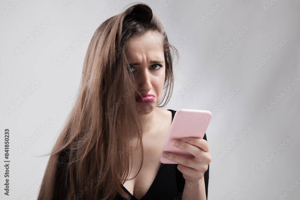 Young woman frowning while looking at her smartphone, expressing confusion and frustration Young woman frowning while looking at her smartphone, expressing confusion and frustration