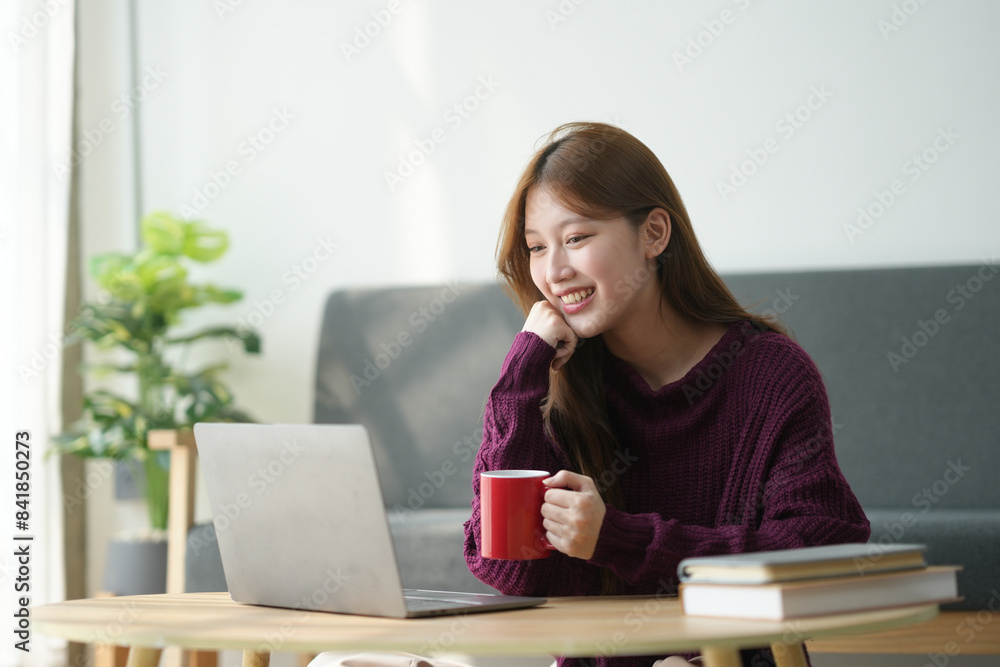Young woman smiling while working on her laptop at home, holding a red coffee mug, seated on a couch with books on the table. Young woman smiling while working on her laptop at home, holding a red coffee mug, seated on a couch with books on the table.