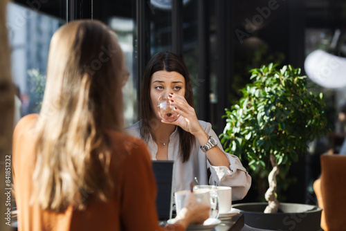 Two young business women in a cafe having one on one meeting. Friends after work talking gossiping and having coffee at a window table with reflections..