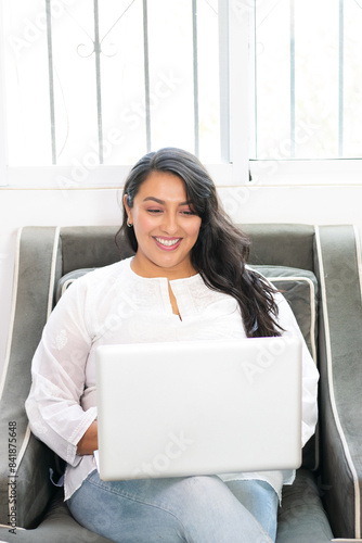 beautiful young mexican woman sitting on sofa in apartment using laptop doing home office, latina working smiling happy