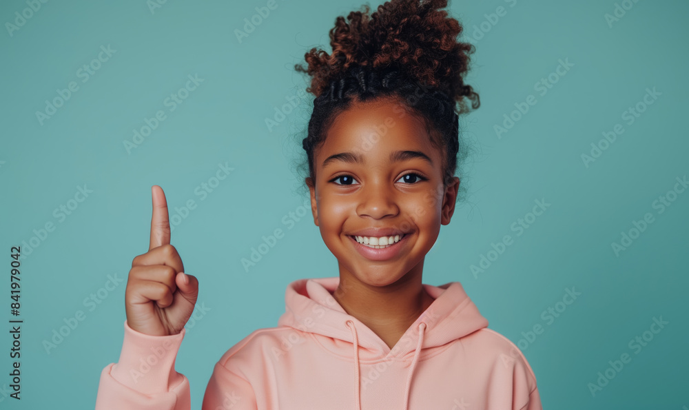 Young african american teenager girl in a pink sweatshirt smiling with healthy teeth and pointing index finger up at copy space expressing wow emotion on a mint background Young african american teenager girl in a pink sweatshirt smiling with healthy teeth and pointing index finger up at copy space expressing wow emotion on a mint background