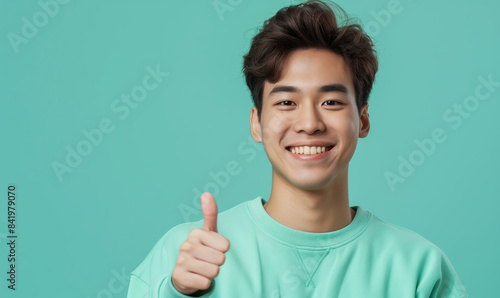 Young asian man in a mint sweatshirt smiling with healthy teeth showing thumb up at copy space expressing wow emotion standing isolated on mint background © anatoliycherkas