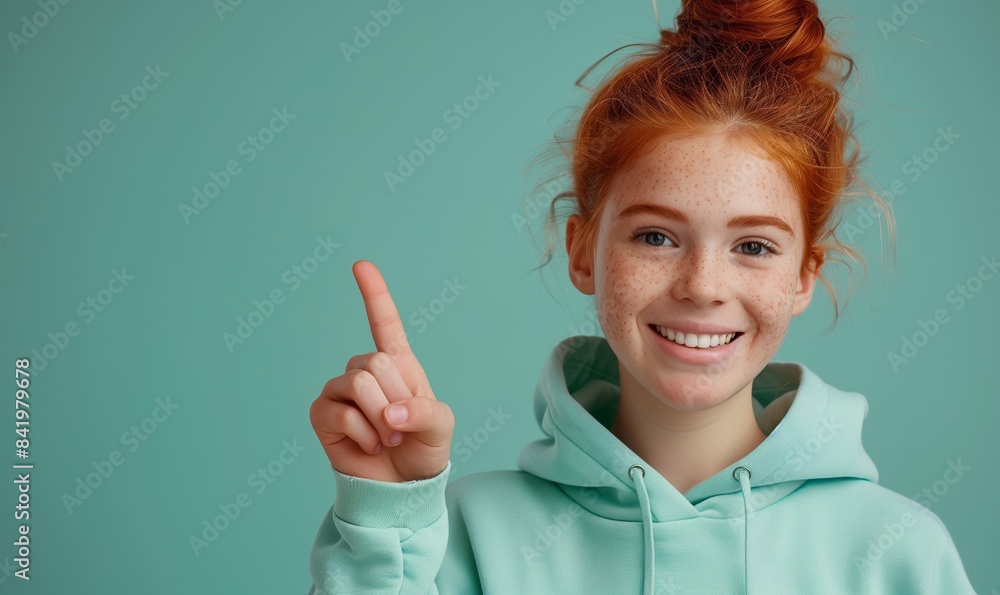 Smiling ginger teenager girl in a mint sweater smiling with healthy teeth and pointing index finger up at copy space expressing wow emotion standing isolated on paper mint background Smiling ginger teenager girl in a mint sweater smiling with healthy teeth and pointing index finger up at copy space expressing wow emotion standing isolated on paper mint background