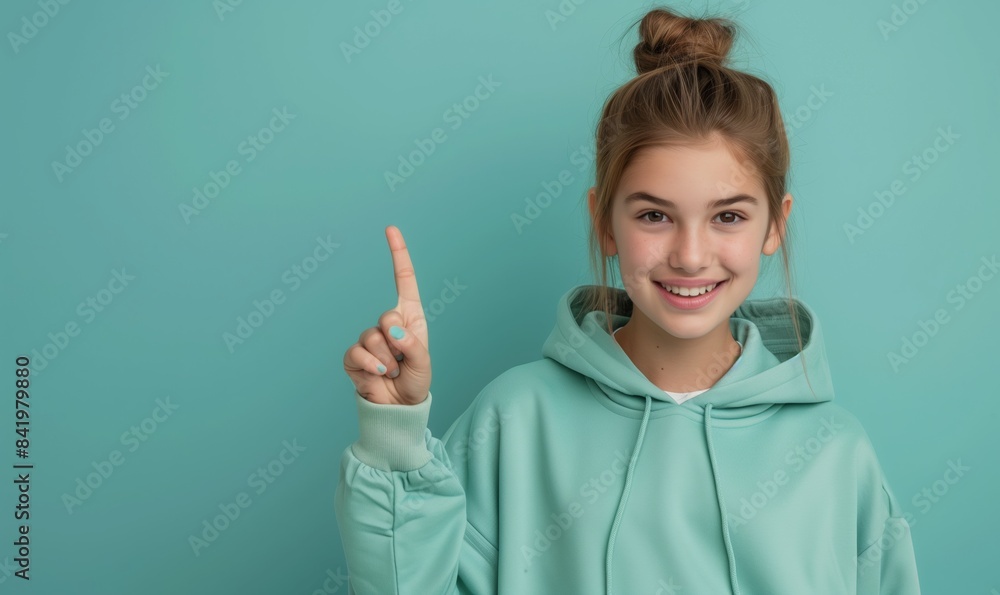Smiling caucasian teenager girl in a mint sweatshirt smiling with healthy teeth and pointing index finger up at copy space expressing wow emotion standing isolated on paper mint background Smiling caucasian teenager girl in a mint sweatshirt smiling with healthy teeth and pointing index finger up at copy space expressing wow emotion standing isolated on paper mint background