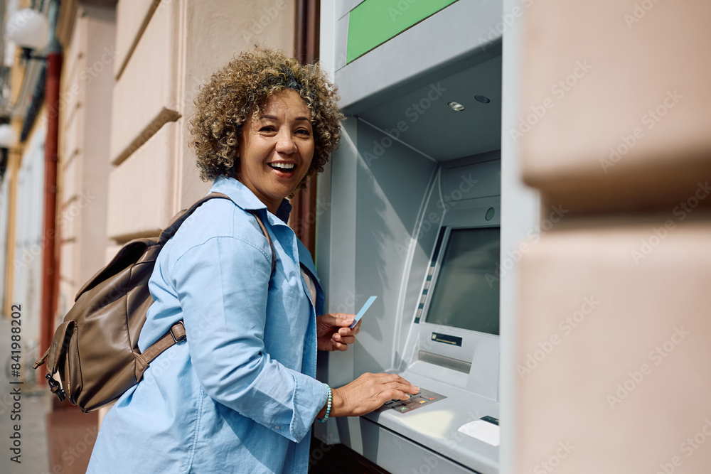 Obraz premium Happy female tourist entering pin while using ATM machine and looking at camera.