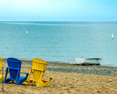 Blue and yellow chairs on a sand beach near a white row boat on the edge of Lake Ontario with a white bouy in the distance room for text