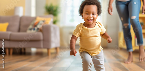 Cute toddler taking his first steps in bright and sunny living room, with hist parent watching in awe in background. photo