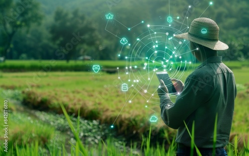 The farmer is using his smartphone to control an agricultural smart farm with digital icons floating above the rice field, on which you can see data about water flow and planting of crops