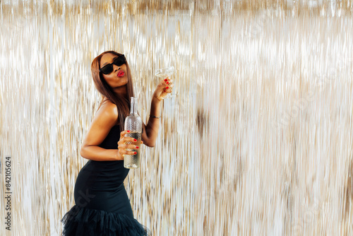 Young happy fashion African girl black woman in cocktail dress and sunglasses holding drink glass and wine bottle dancing at party with glitter background having fun at holiday, birthday celebration.