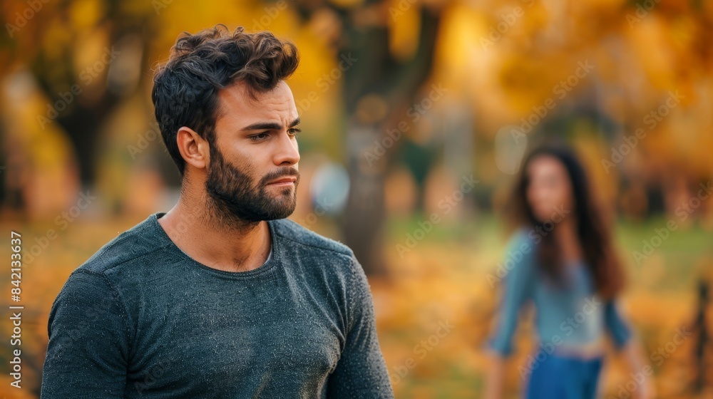 Sad young man with beard in park during autumn, with woman in background. Concept of break-up and loneliness. Sad young man with beard in park during autumn, with woman in background. Concept of break-up and loneliness.