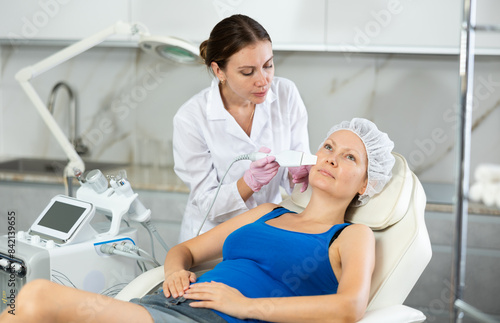 Young female cosmetologist performs facial exfoliating procedure with machine to adult female client
