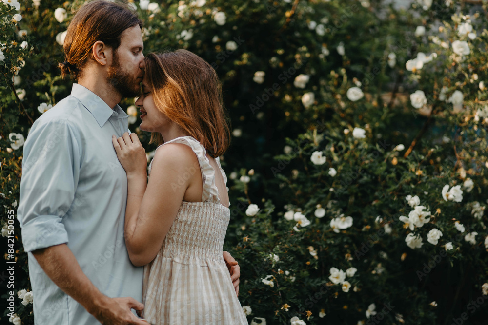 Fototapeta premium beautiful couple in retro clothes hugging near a tree of white roses in the summer in the village.