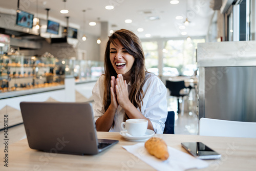 Beautiful and happy young woman sitting and eating delicious rolls in bakery or fast food while she using laptop computer.