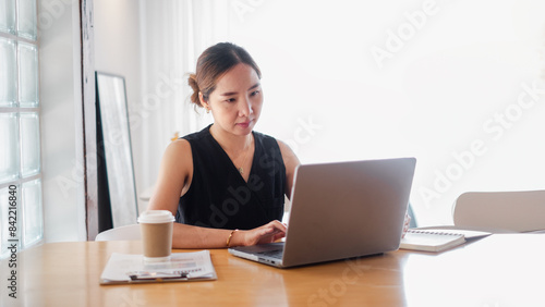 A woman is sitting at a desk with a laptop and a cup of coffee. She is focused on her work and she is in a serious mood © Mongta Studio