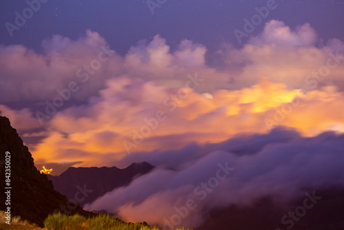 Under the veil of night, the dramatic clouds above Pico do Arieiro dance in a mesmerizing display, illuminated by the vibrant hues of magenta and orange city lights below.