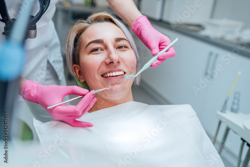 Portrait of young smiling woman sitting in stomatology clinic chair and doctor's hands with dental Mouth mirror and Dental explorer tools preparing to examine teeth condition. Health care concept.