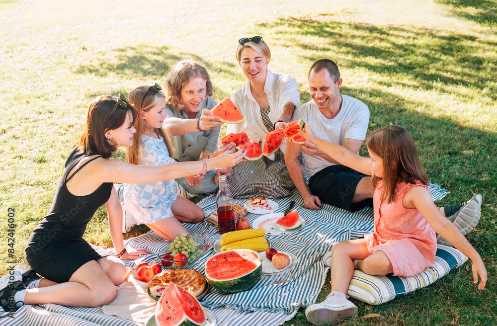 Big caucasian family on picnic blanket on the in city park green grass. They rose-up red juicy watermelon pieces and clinking them like glasses. Family values and outdoor activities concept. Big caucasian family on picnic blanket on the in city park green grass. They rose-up red juicy watermelon pieces and clinking them like glasses. Family values and outdoor activities concept.