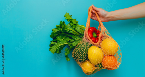 A hand holding an orange net bag with vegetables and fruits on a blue background. photo