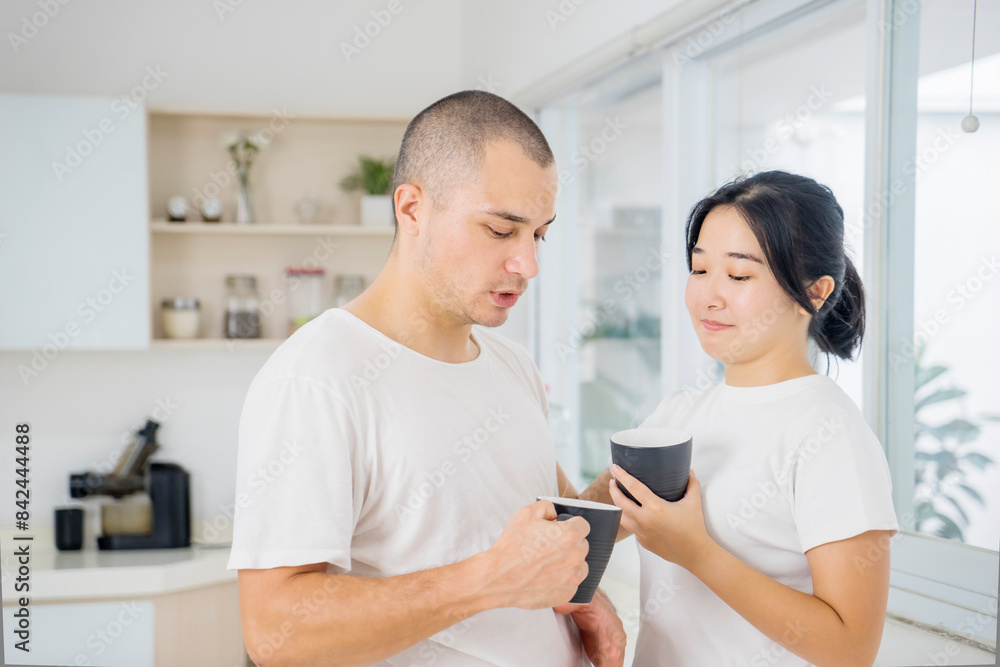 Portrait shot of cute smiling young Asian lover couple standing near the windows in the living room in morning, holding and drinking cup of coffee while looking at each other eyes and talking with fun Portrait shot of cute smiling young Asian lover couple standing near the windows in the living room in morning, holding and drinking cup of coffee while looking at each other eyes and talking with fun