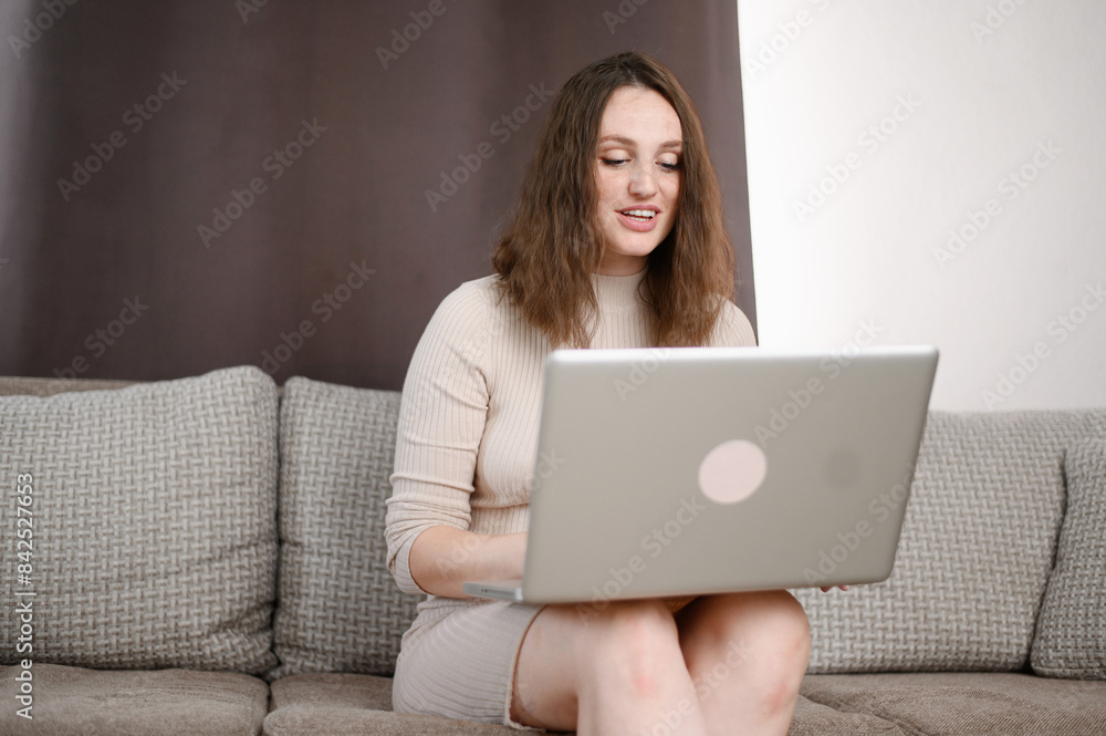 Smiling young woman in dress working at home, sitting on sofa, using laptop. Female student studying online, browsing web and typing on computer, watching videos or studying electronically. Smiling young woman in dress working at home, sitting on sofa, using laptop. Female student studying online, browsing web and typing on computer, watching videos or studying electronically.