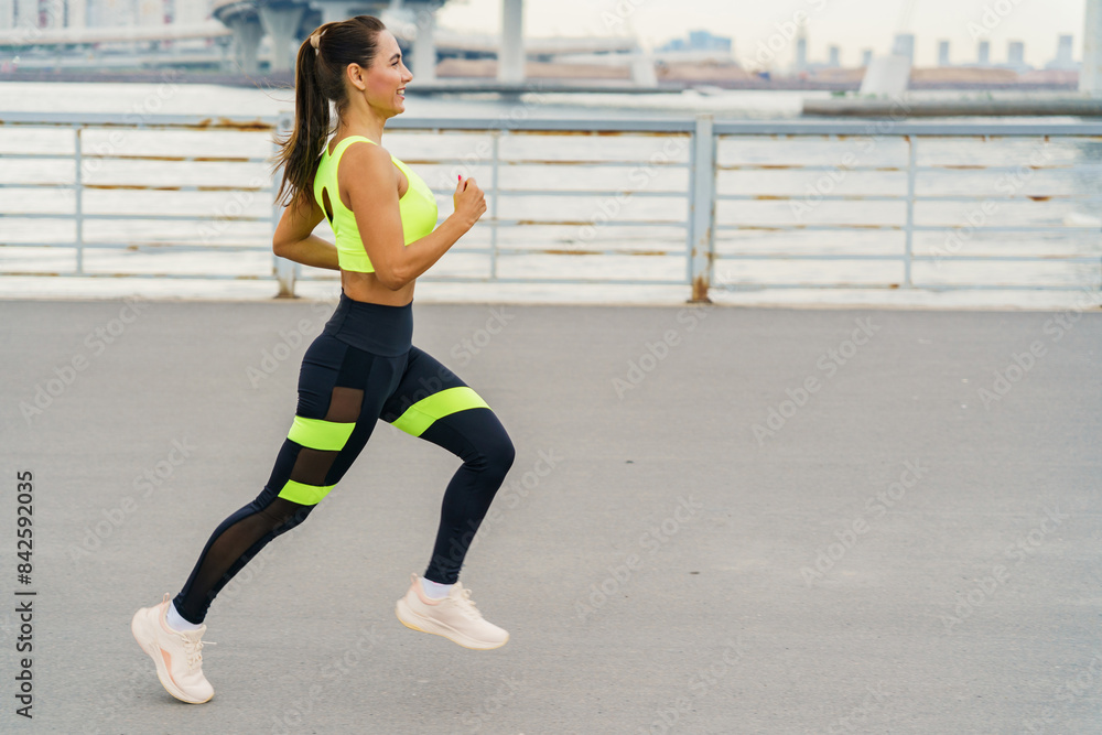 A woman jogs along an urban waterfront, smiling confidently in bright sportswear, embodying fitness and joy. A woman jogs along an urban waterfront, smiling confidently in bright sportswear, embodying fitness and joy.