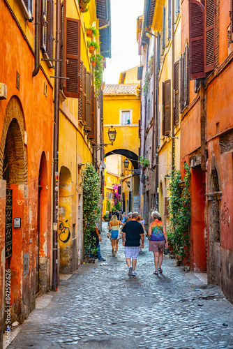 Fototapeta Naklejka Na Ścianę i Meble -  Tourists walk through a narrow, cobblestone alley in Rome, Italy. The buildings are brightly colored and adorned with plants and flowers. The alley leads to an archway, providing a glimpse of the city