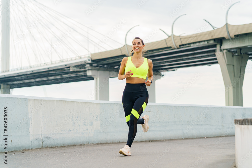 A woman jogs along a waterfront path, smiling confidently in vibrant sportswear, with a modern bridge in the background. A woman jogs along a waterfront path, smiling confidently in vibrant sportswear, with a modern bridge in the background.