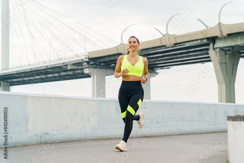 A woman jogs along a waterfront path, smiling confidently in vibrant sportswear, with a modern bridge in the background. © muse studio