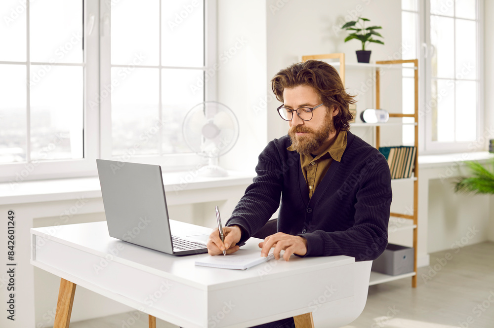 Busy man working in the office. Serious bearded businessman in eyeglasses writing something on a notepad while sitting at his white desk with a laptop computer Busy man working in the office. Serious bearded businessman in eyeglasses writing something on a notepad while sitting at his white desk with a laptop computer