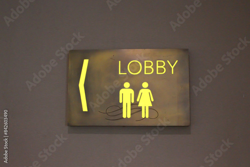 The guide points to the toilet with a yellow wooden background,  lobby sign with gold color text photo taken in cinema
