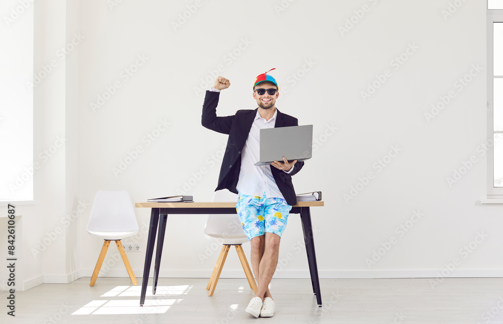 Funny happy business man standing on workplace in office wearing beach shorts and suit jacket holding laptop and booking tickets for summer vacation online. Travel, vacation and holiday concept. Funny happy business man standing on workplace in office wearing beach shorts and suit jacket holding laptop and booking tickets for summer vacation online. Travel, vacation and holiday concept.