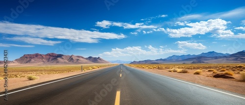 A long road stretches across a desert landscape. The sky is clear and blue, and the sun is shining brightly. The road is wide and empty, with no cars or other vehicles in sight. The scene is peaceful