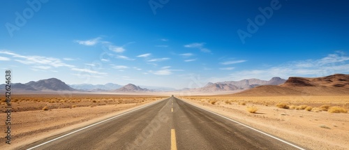A long road stretches across a desert landscape. The sky is clear and blue, and the sun is shining brightly. The road is wide and empty, with no cars or other vehicles in sight. The scene is peaceful